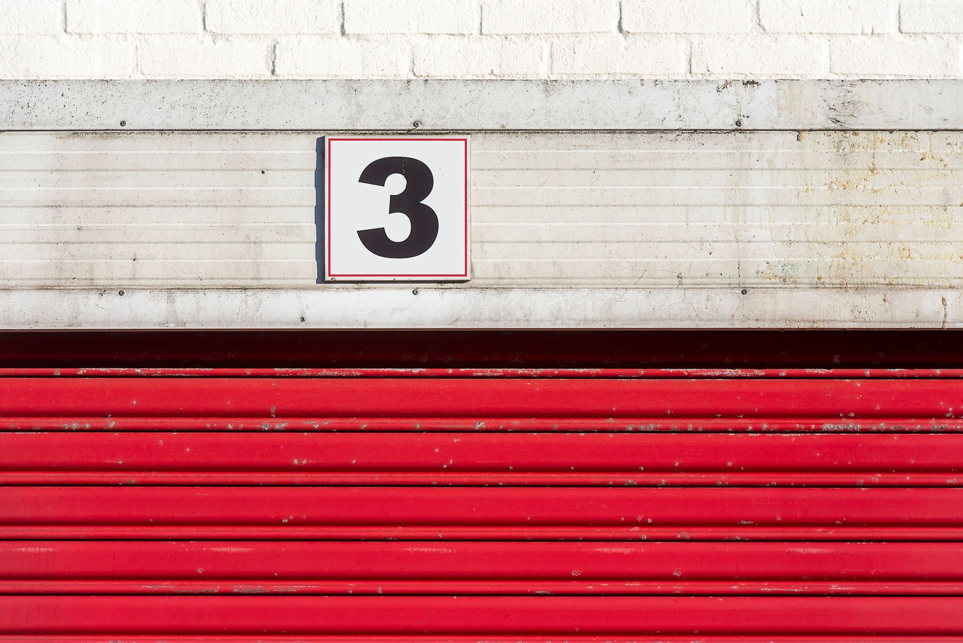 Red garage door with a white sign marked “3” on a light brick wall.