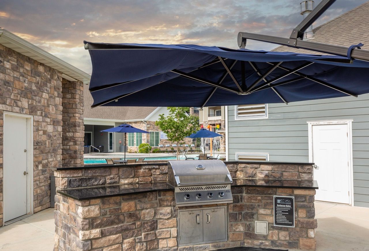 Outdoor community grilling station with a stone counter, blue umbrellas, and a pool in the background at Herriman Towne Center in Herriman, UT.