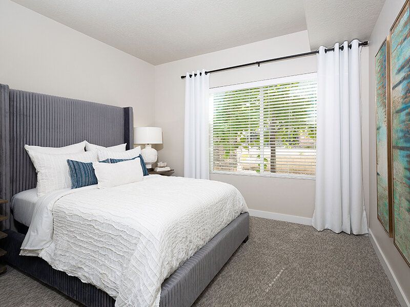 Bedroom in a rental unit with a gray upholstered bed, white bedding, and a window with white curtains at Herriman Towne Center in Herriman, UT.