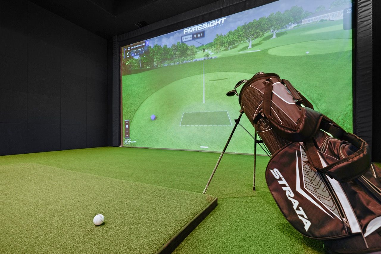 Indoor golf simulator room with turf, a golf bag, and a large screen showing a golf course at Herriman Towne Center in Herriman, UT.
