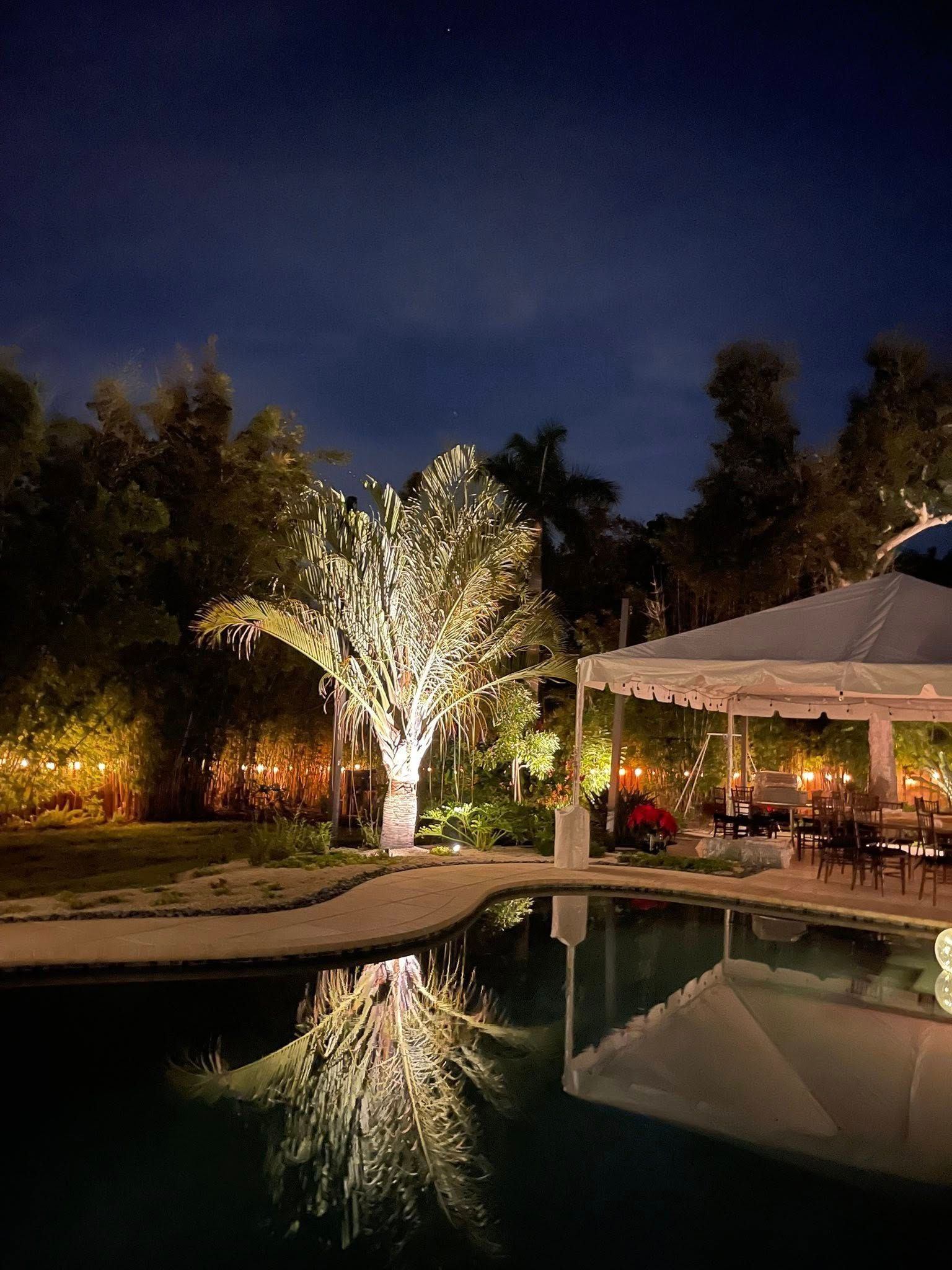 Nighttime poolside garden with lit trees and a pavilion reflected in calm water