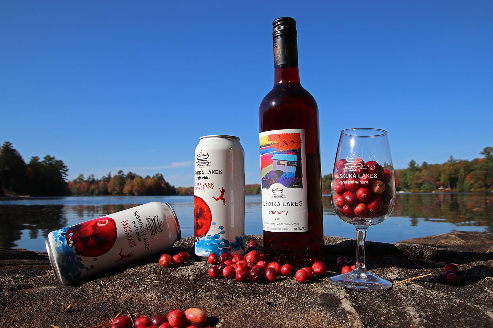 A bottle of wine , two cans of beer , and a glass of wine are sitting on a rock near a lake.