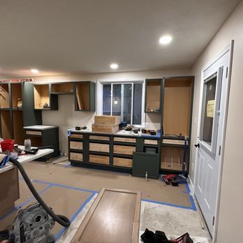 Kitchen cabinets being installed; dark green lower cabinets, light upper cabinets, unfinished.