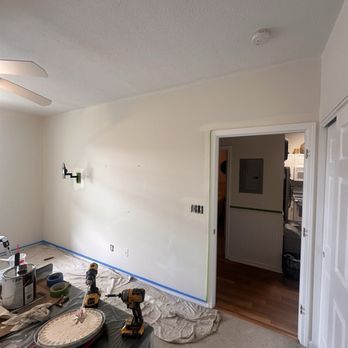 Room being painted, cream walls with painter's tape, tools, doorway to another room, white door.