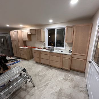 Kitchen under construction with light wood cabinets, countertop, and window. Gray flooring, tools, and a ladder are present.