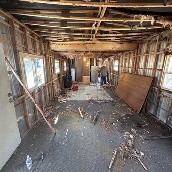 Interior of a gutted building with exposed wood framing. A person stands in the center. Debris litters the floor.
