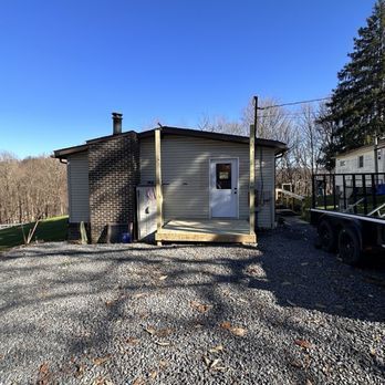 Small, beige building with a wooden porch and a brick chimney on a gravel lot under a blue sky.