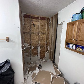 Bathroom wall under construction, drywall removed, revealing wood frame. Debris on floor; wood cabinets.