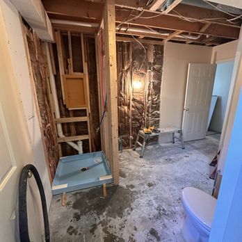 Bathroom under construction, showing shower base, partially framed walls, and exposed pipes/wiring.