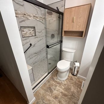 Bathroom with marble-tiled shower, glass door, toilet, and wooden cabinet. Tan and white tones.