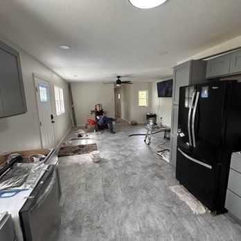 Interior view of a kitchen under renovation, with a person seated. Gray cabinets, black refrigerator, and gray flooring are present.