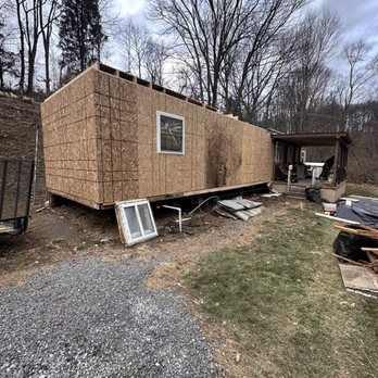 House under construction; exterior walls covered in OSB, porch visible. Grassy area, cloudy sky.
