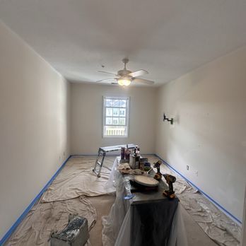 Room with beige walls, a window, ceiling fan, and painting supplies on a covered table.