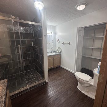 Bathroom with shower, vanity, toilet, and shelving. Dark wood-look floor, light walls, and open shower.