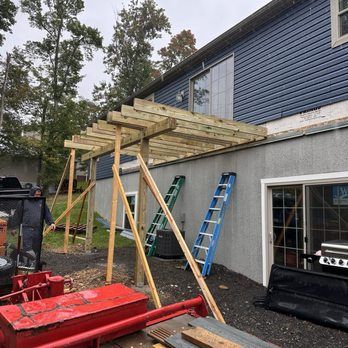 Construction of a wooden pergola attached to a house. Two ladders lean against the wall. A person is nearby.