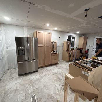 Kitchen under construction with cabinets and stainless steel refrigerator. Man standing in the room. Gray floors and drywall walls.