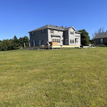 Gray two-story house with a large deck on a grassy hill under a blue sky.