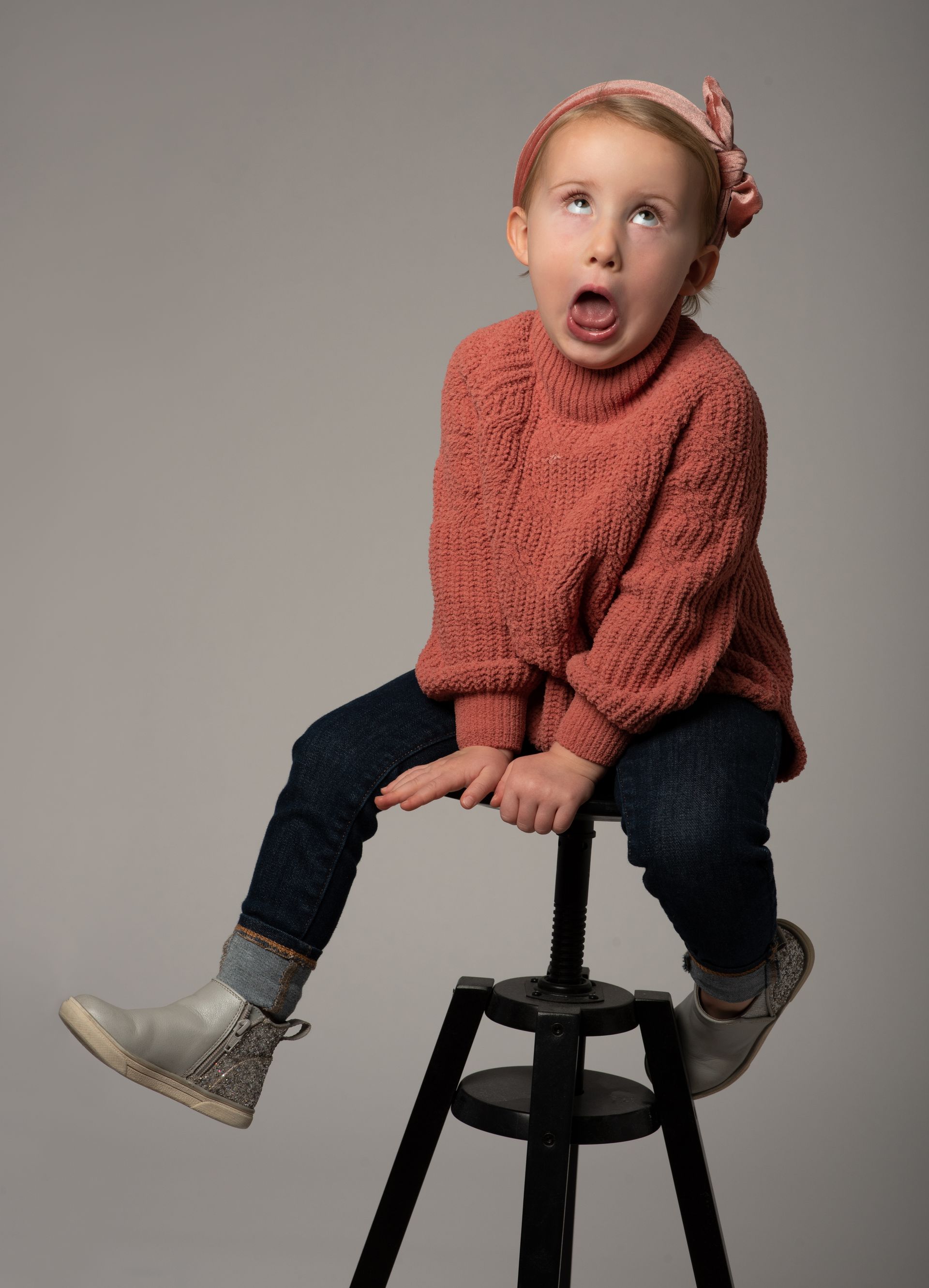 Child sitting on a stool, wearing a pink sweater and dark pants, with a surprised expression