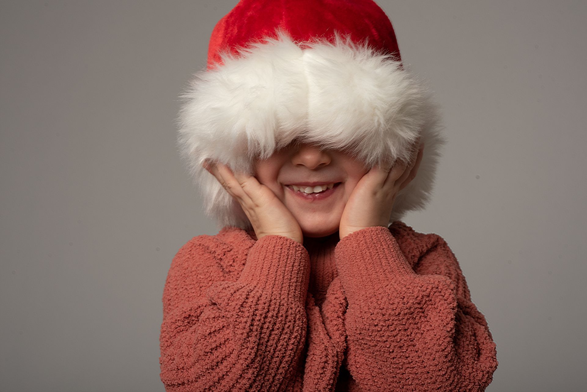 Child in a red Santa hat with white fur brim, smiling and covering their face with hands.