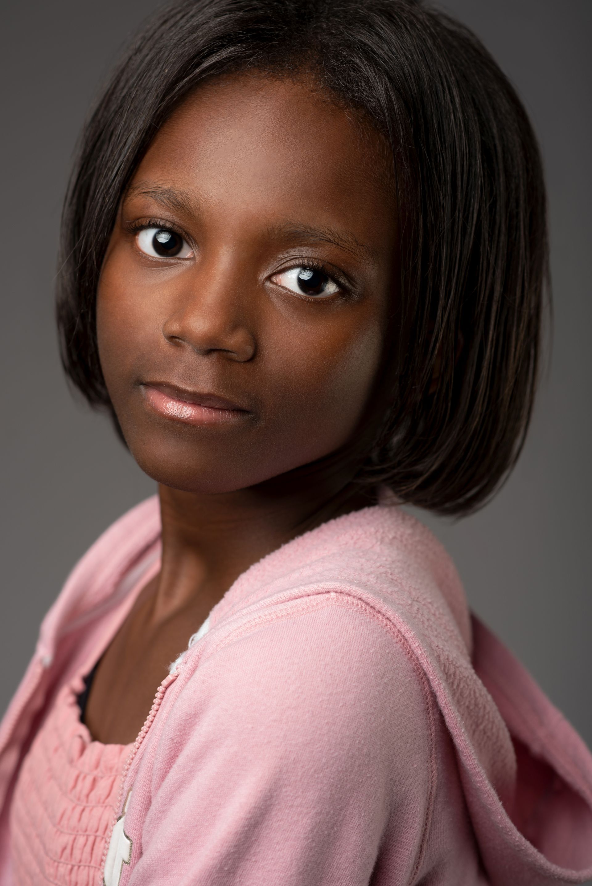 Portrait of a young person with short dark hair wearing a pink top, looking at the camera against a gray background