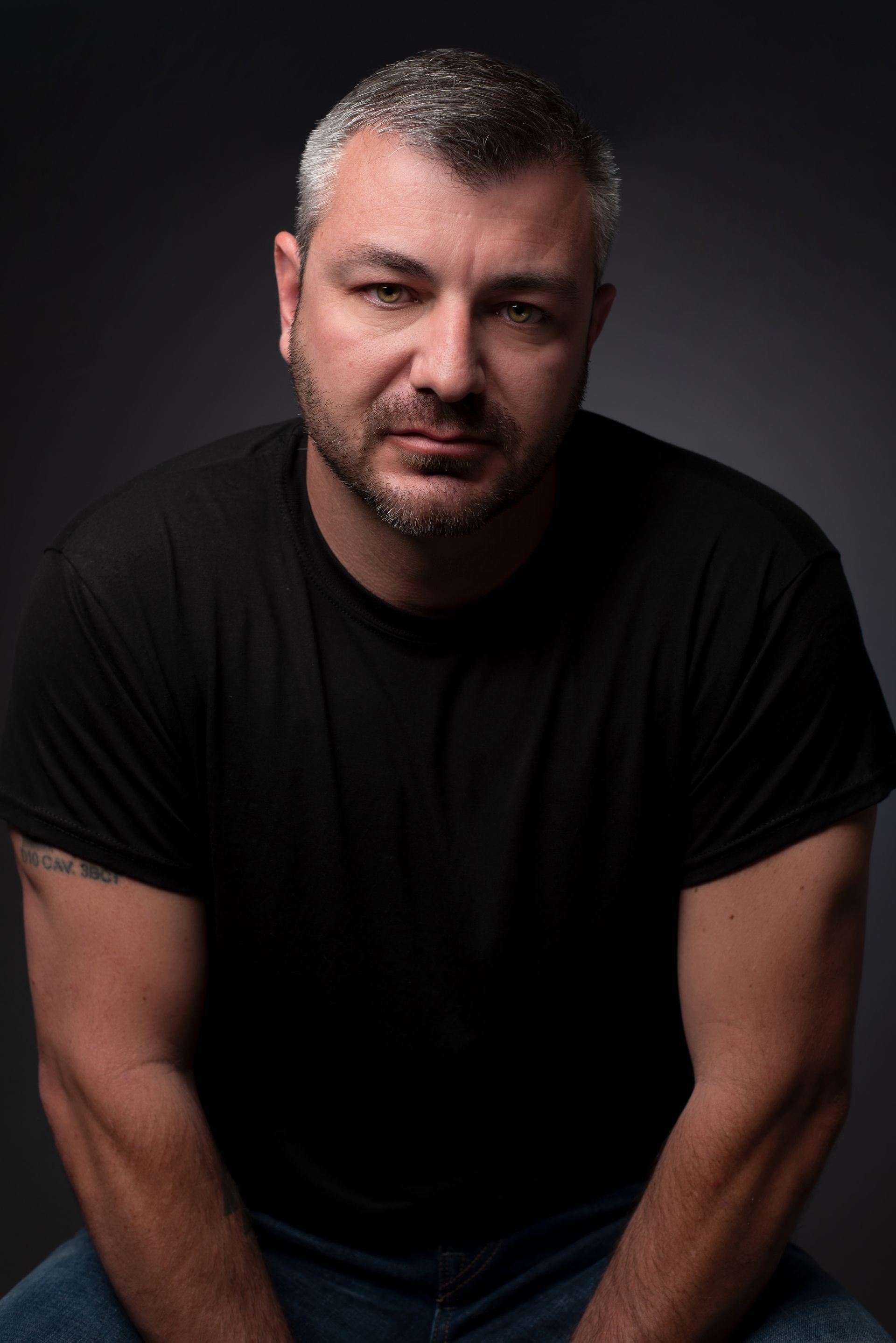 Man in a black T-shirt seated against a dark background, looking at the camera.