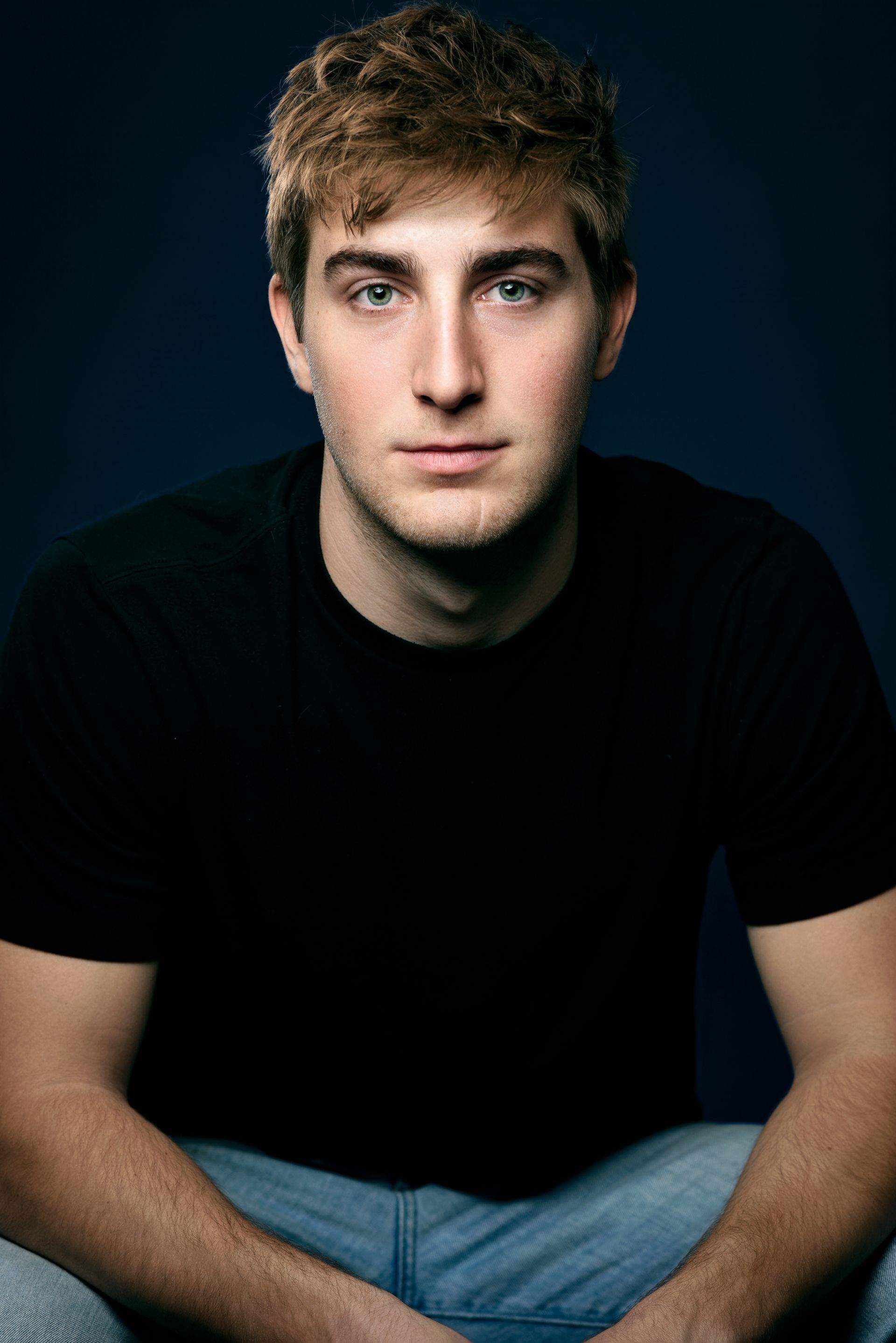 Portrait of a person in a black shirt and jeans, seated against a dark background, looking at the camera.