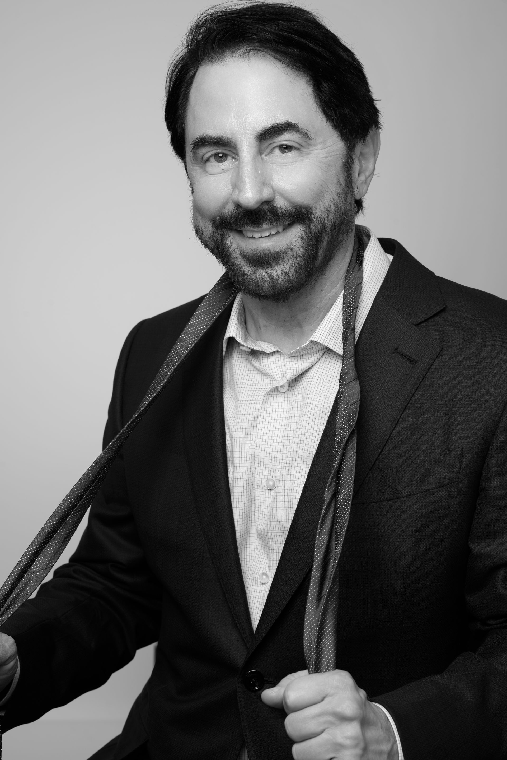 Man in a dark blazer holding a guitar, smiling against a plain gray background.