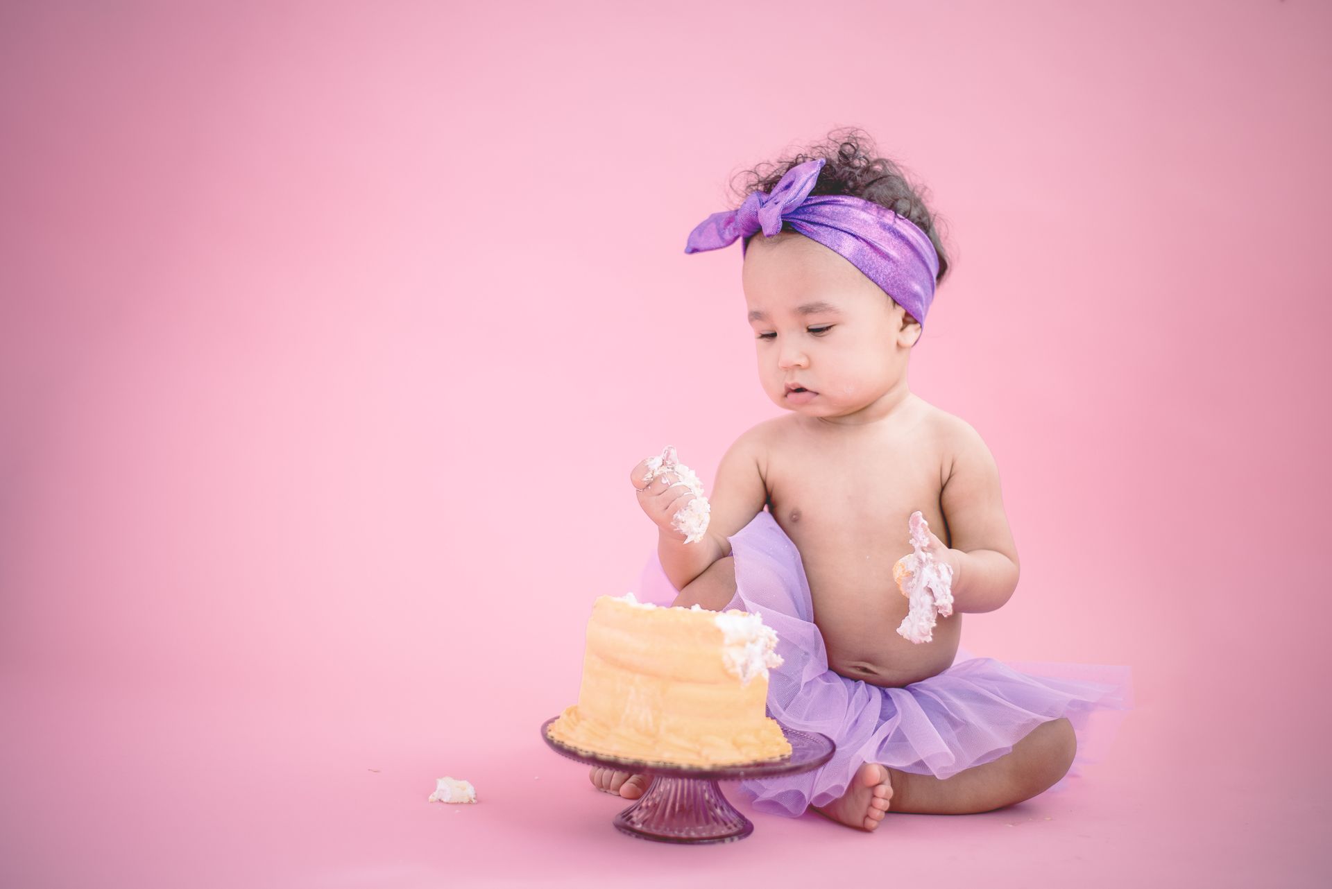 Baby in a purple tutu beside a small frosted cake on a pink backdrop