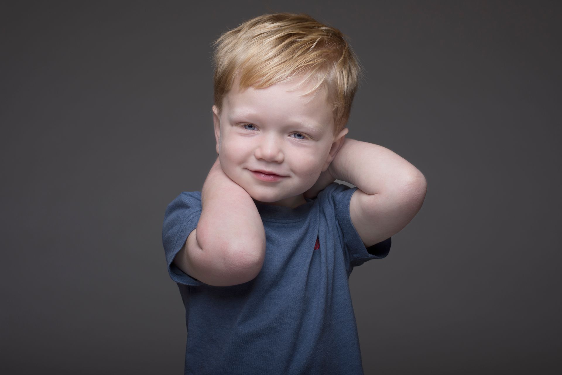 Smiling child in a blue shirt with hands behind their head against a gray background