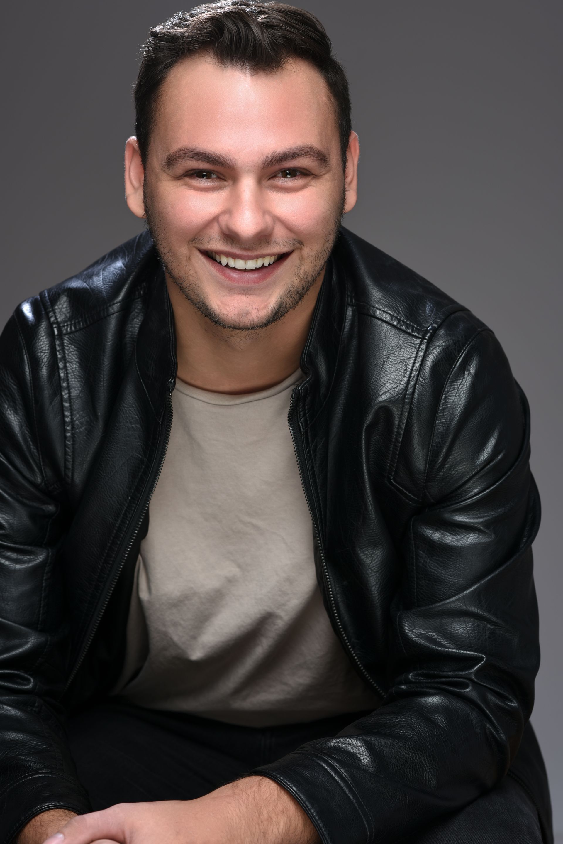 Smiling person in a black leather jacket and beige shirt, seated against a gray background.