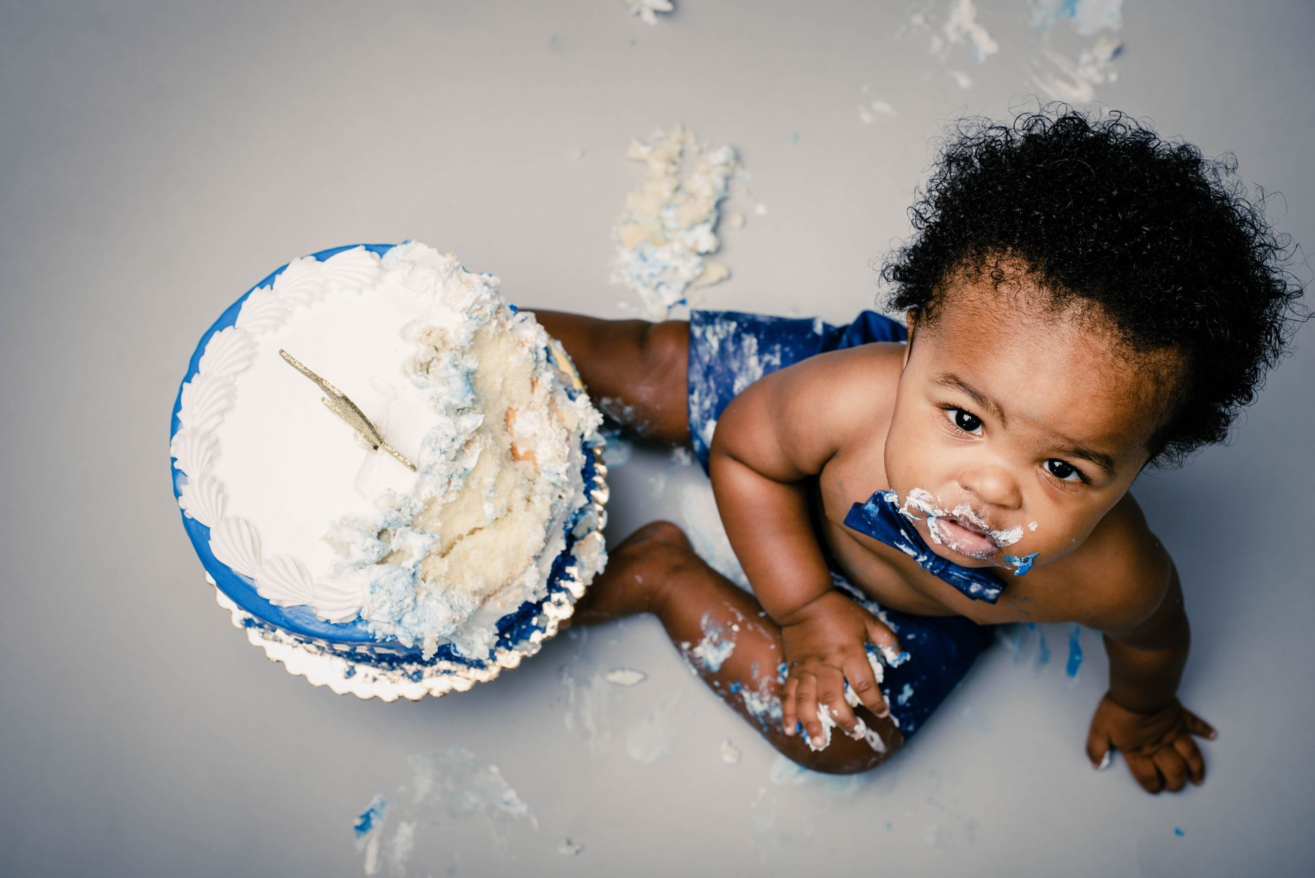 Toddler covered in cake and frosting beside a smashed blue-and-white cake on a gray surface