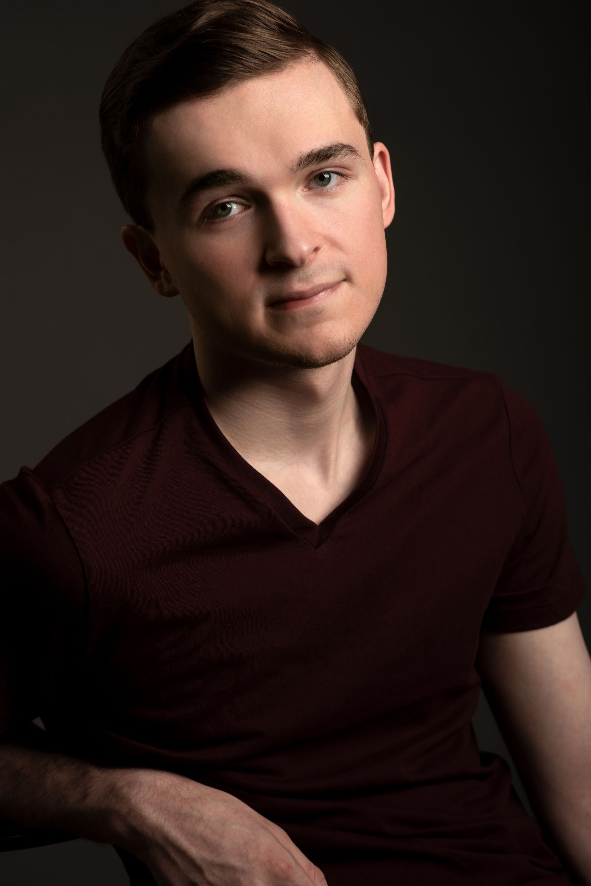 Young man in a dark burgundy shirt posing against a dark studio background.