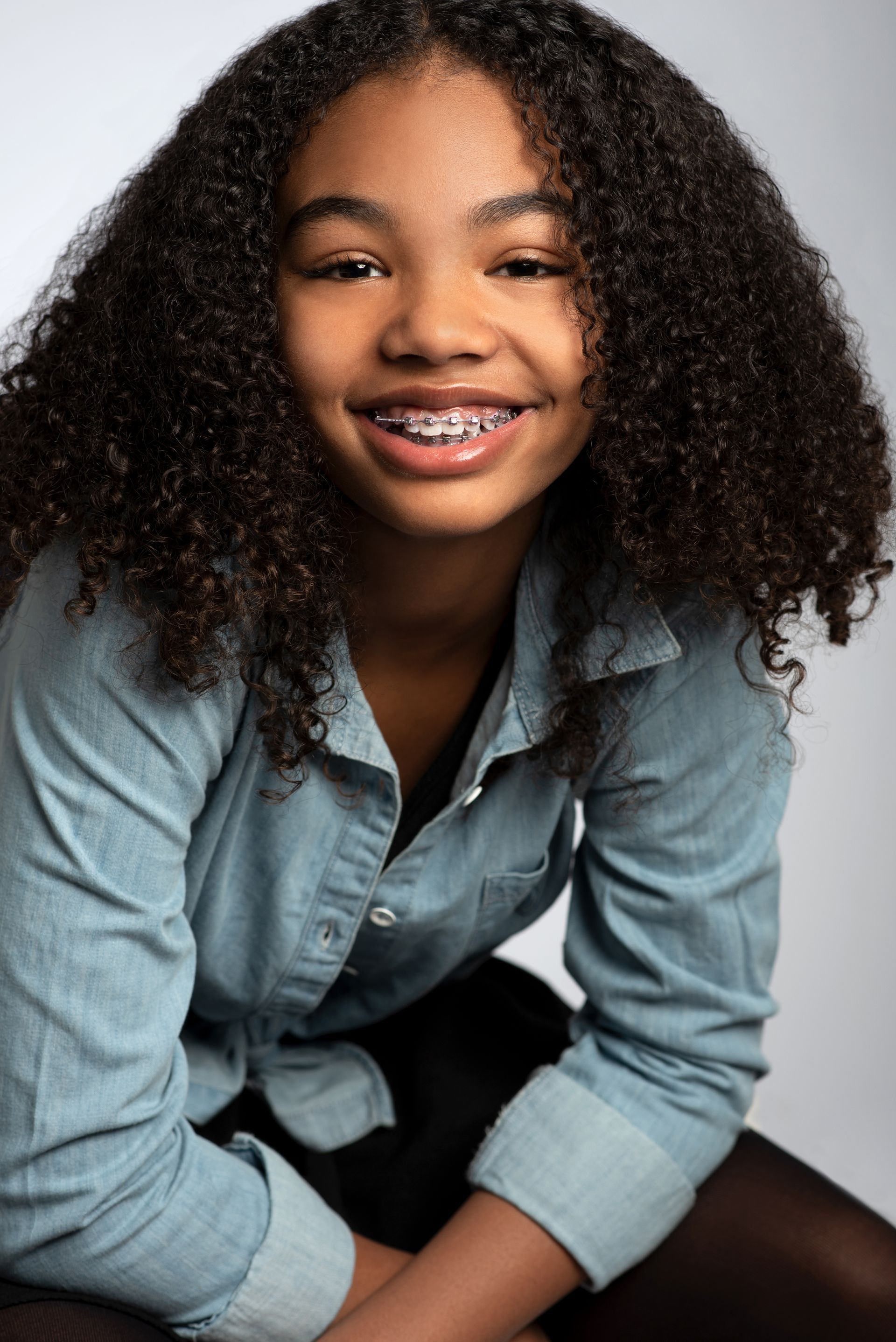 Smiling person with curly hair in a light denim shirt, seated against a plain light background.