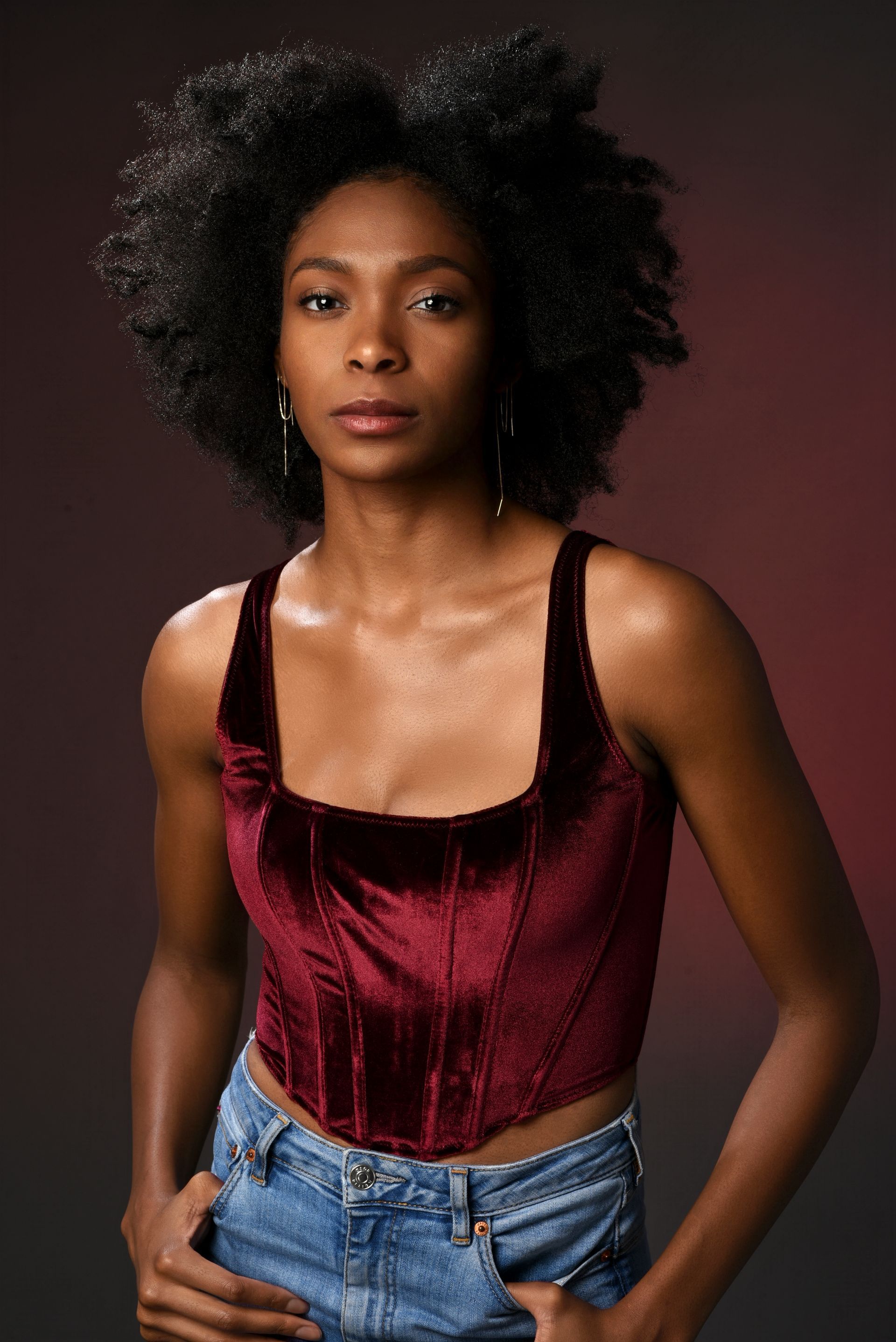 Woman standing against a dark backdrop, wearing a burgundy velvet top and jeans, with a neutral expression.