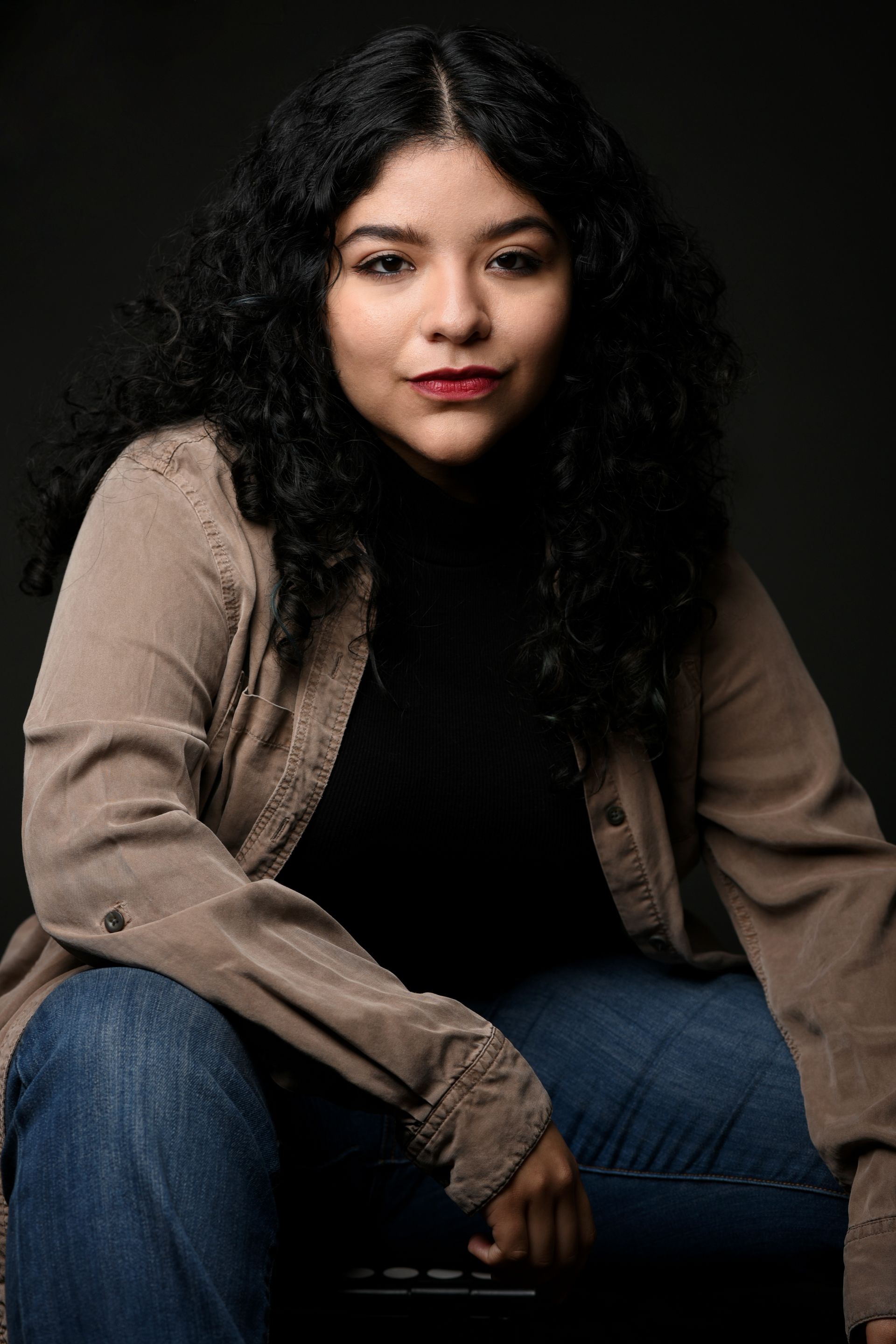 Seated person with curly hair wearing a beige cardigan and jeans against a dark background
