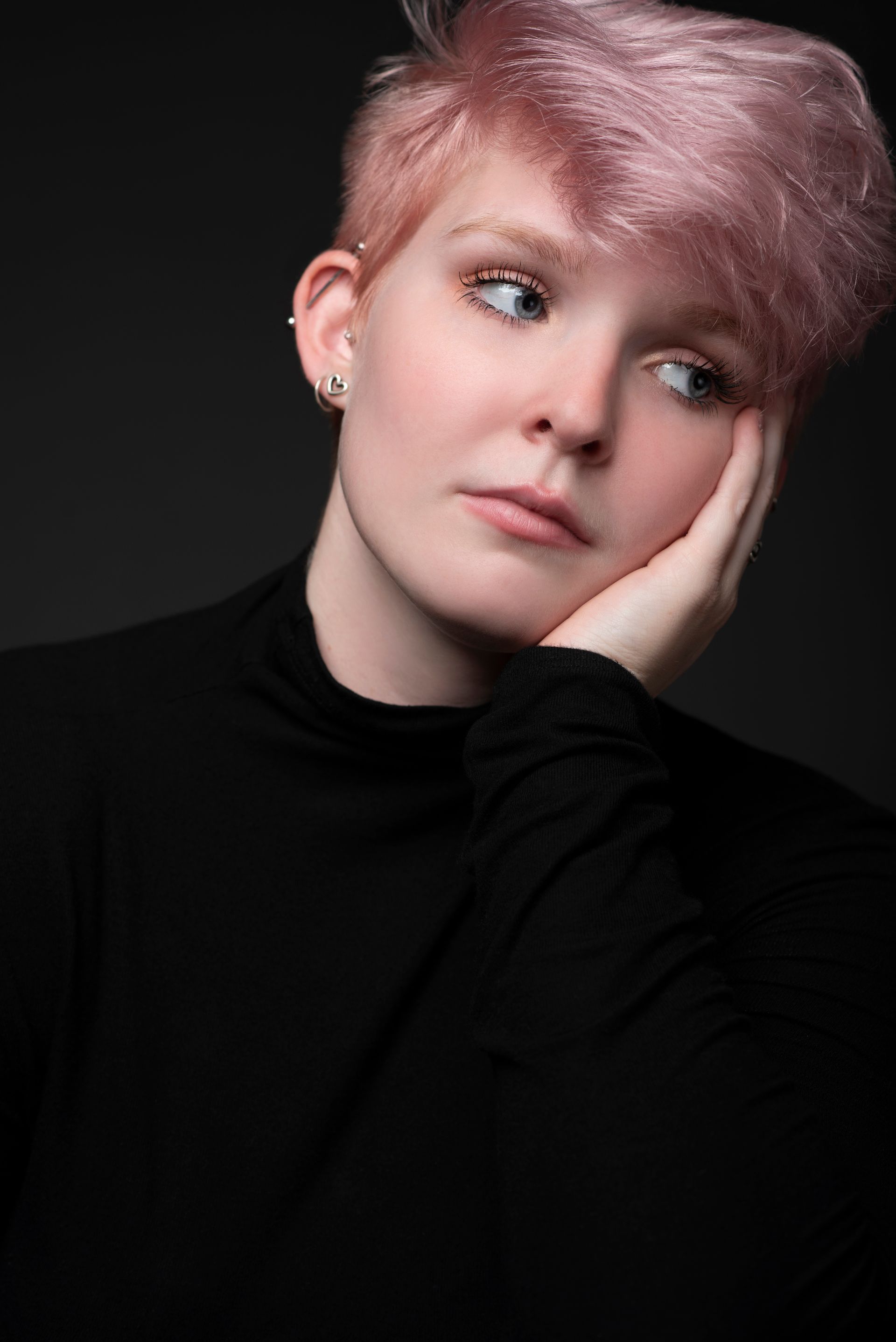 Portrait of a person with short pink hair, resting head on hand, looking off to the side against a dark background