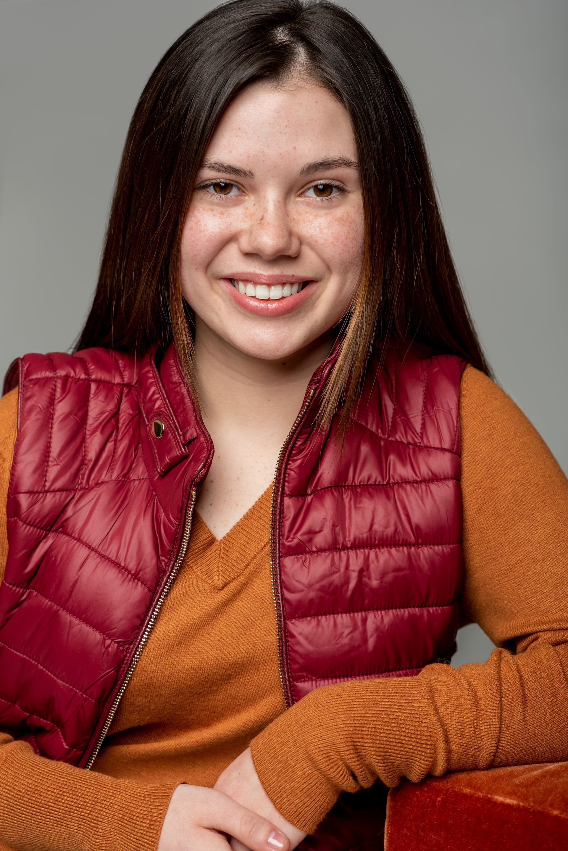 Smiling woman in a red quilted vest and orange sweater, seated against a gray background.