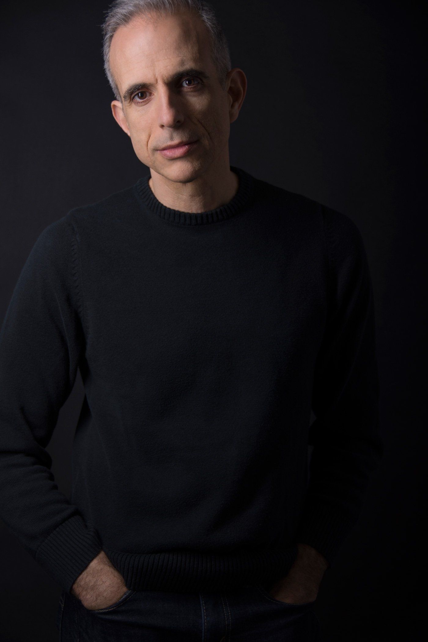 Studio portrait of a man in a black sweater against a dark background, with one hand in his pocket