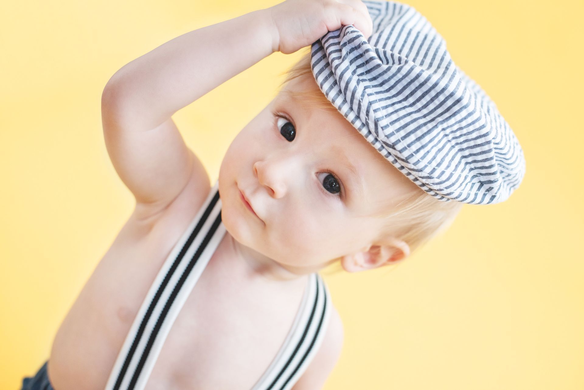 Baby in striped cap and suspenders against a yellow background, tilting head and holding hat