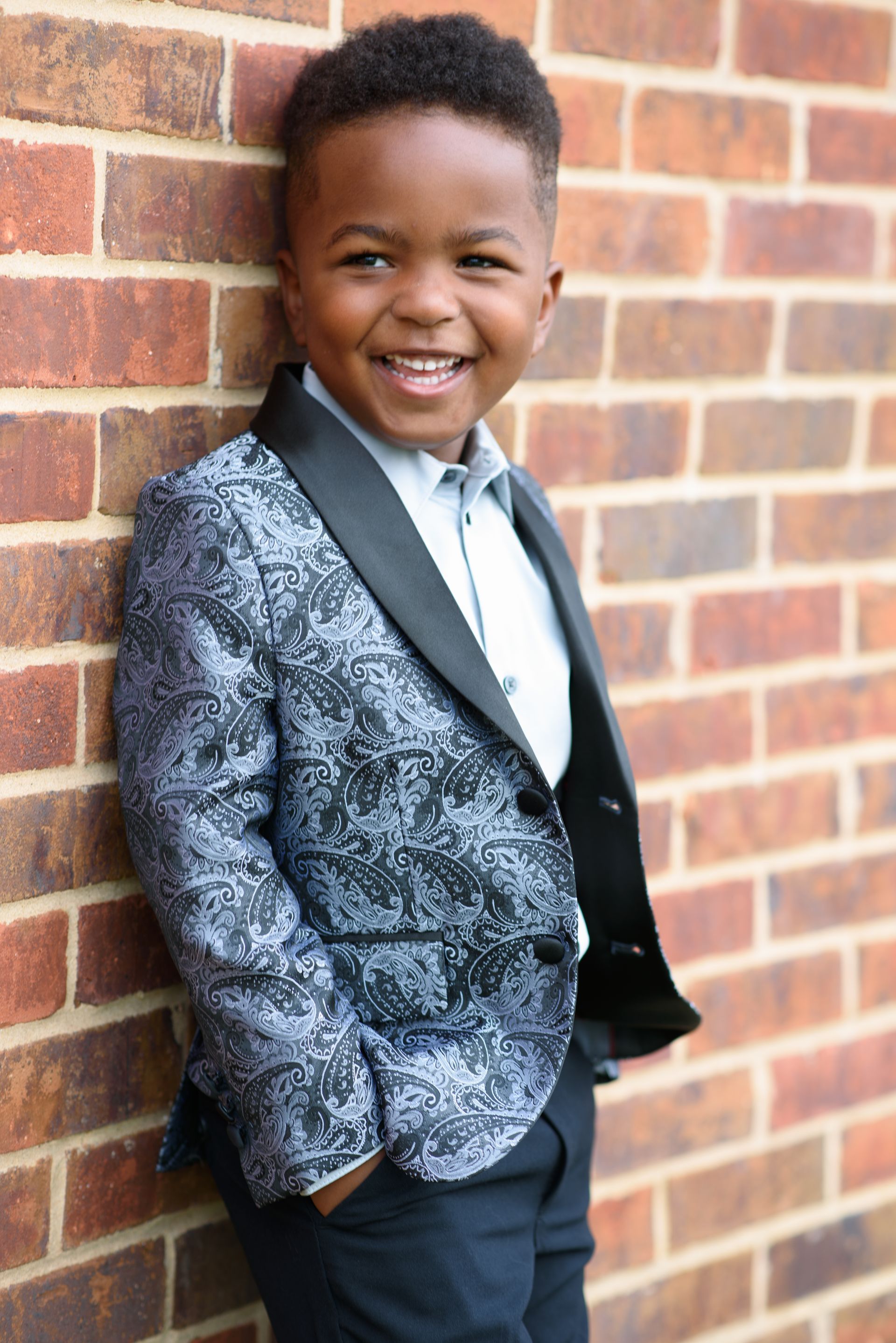 Smiling child in a patterned blazer leaning against a brick wall