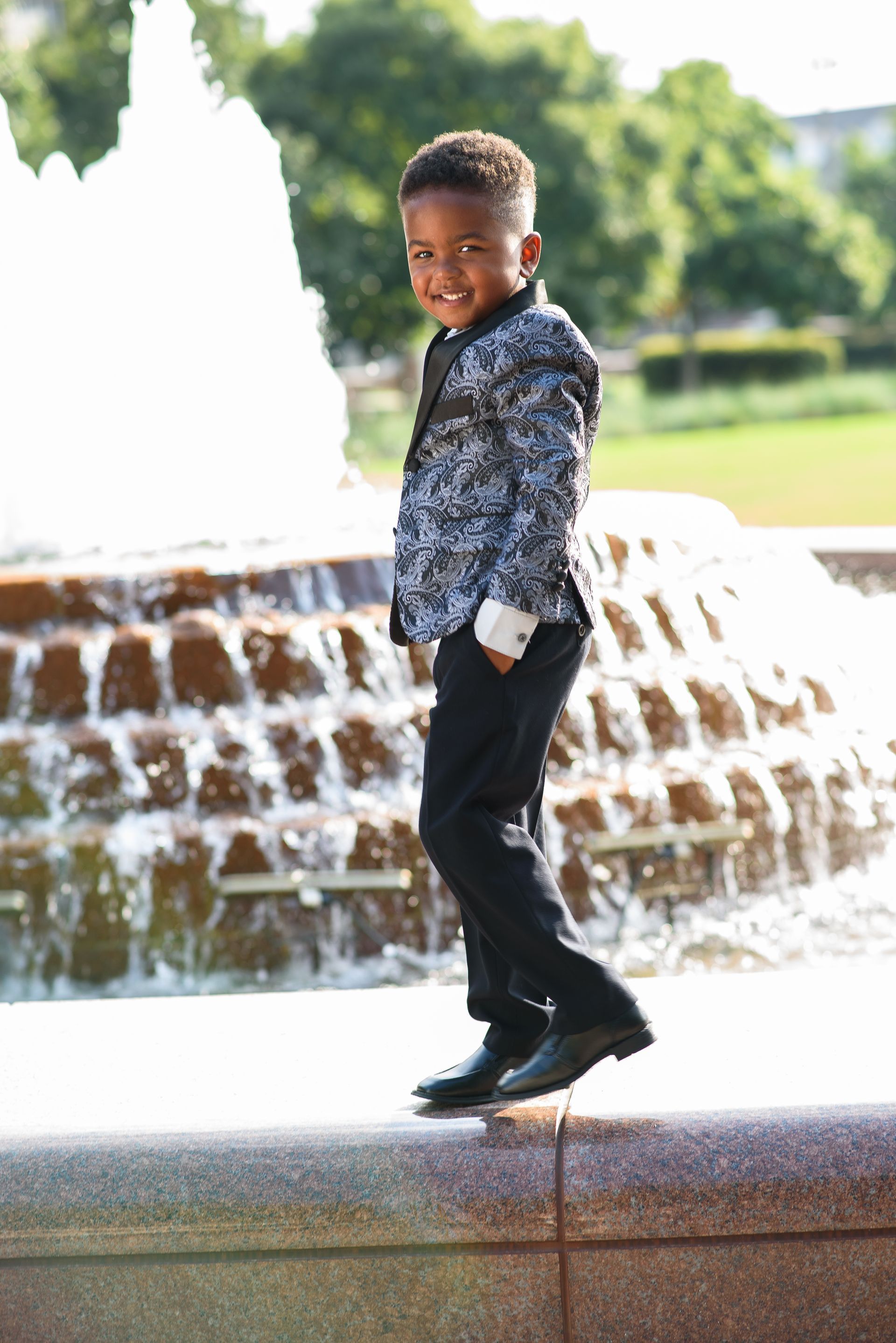 Child posing in front of a fountain, wearing a patterned jacket and black pants.
