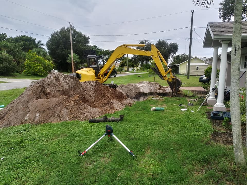 Yellow excavator digging a dirt pile beside a house on a grassy roadside, with a tripod in the foreground