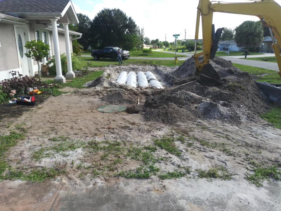 Construction site beside a house with dirt piles, white pipes, and an excavator in a suburban yard