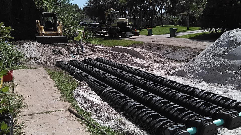Construction site with black drainage pipes stacked in a trench beside gravel piles and machinery