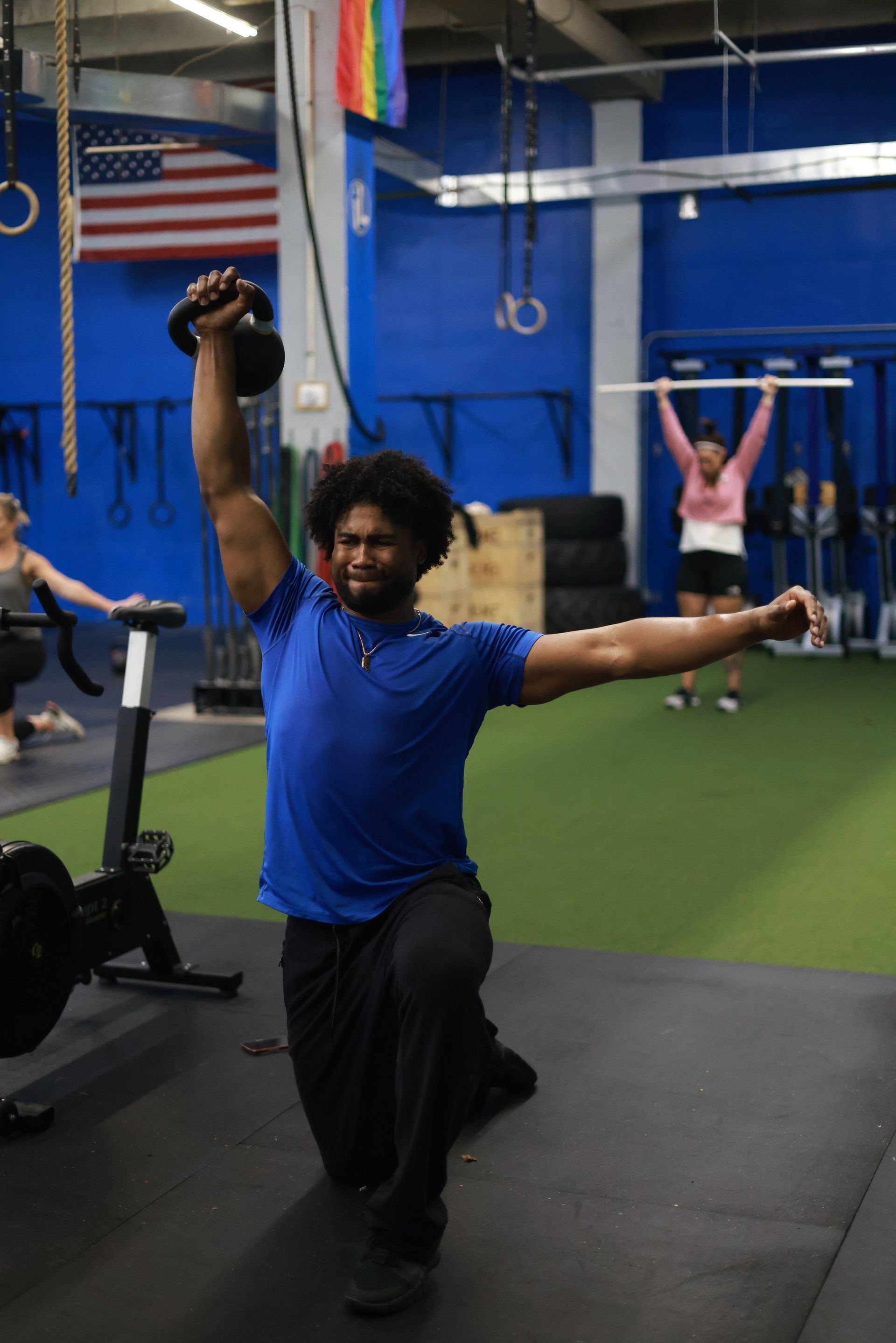 A man is kneeling down in a gym holding a kettlebell over his head.