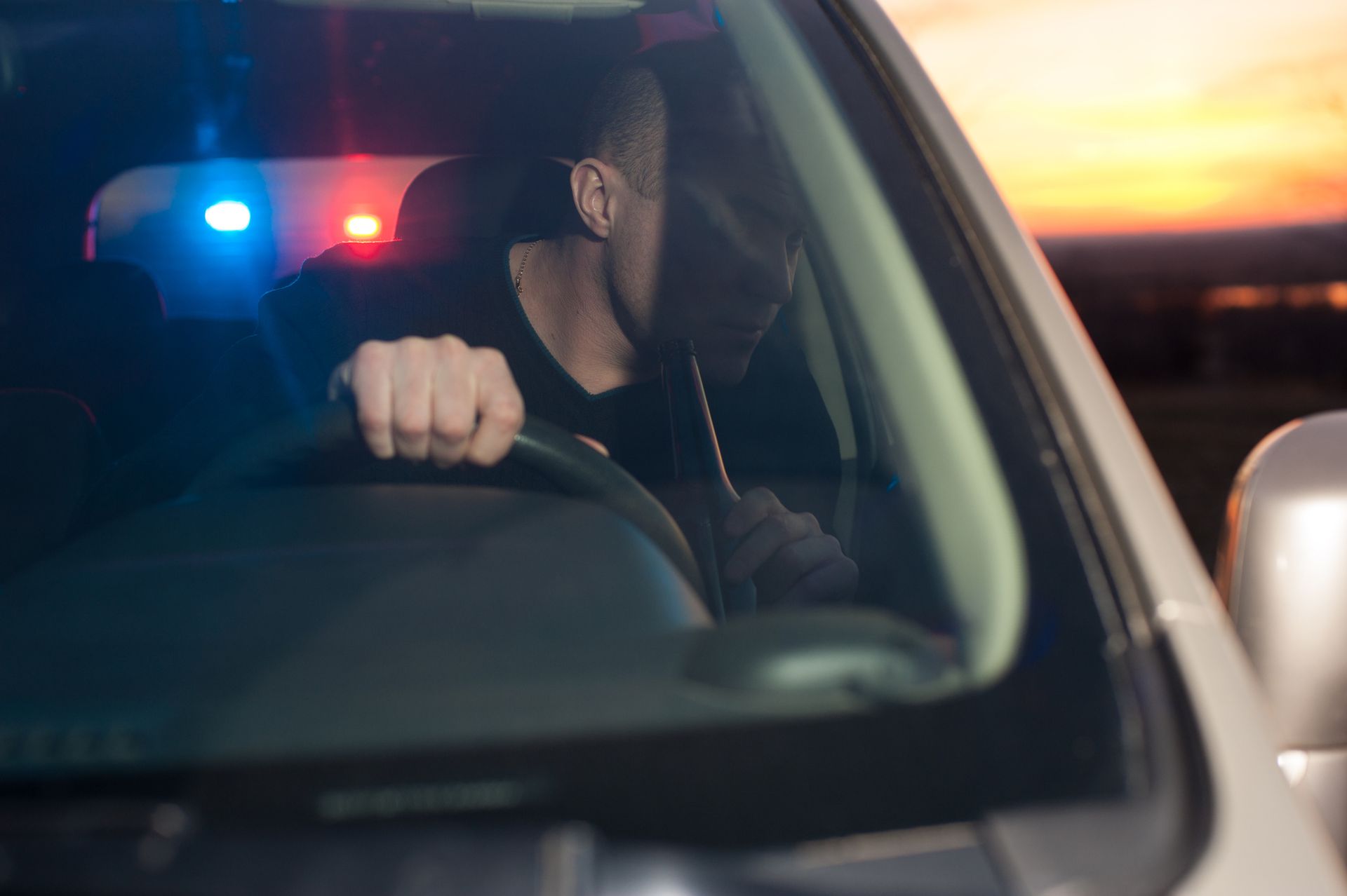 a man driving and holding a beer bottle, with police lights behind him