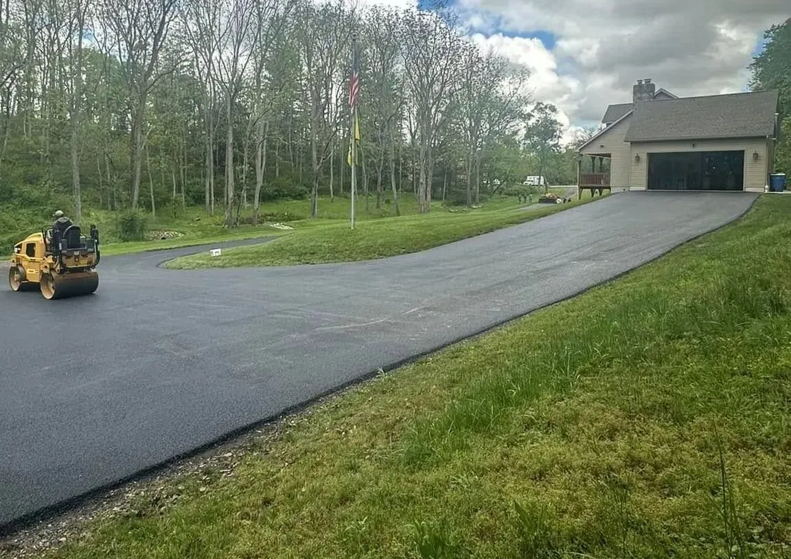 Freshly paved driveway leading to a house beside a grassy lawn and trees, with a roller on the left