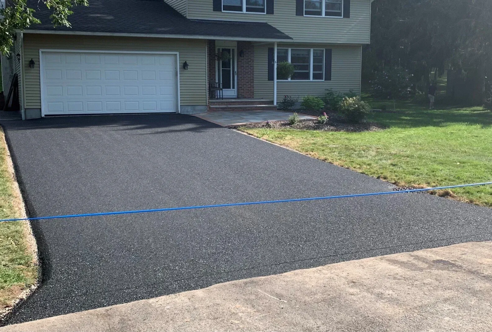 Fresh black asphalt driveway leading to a white garage and two-story house with a small front lawn