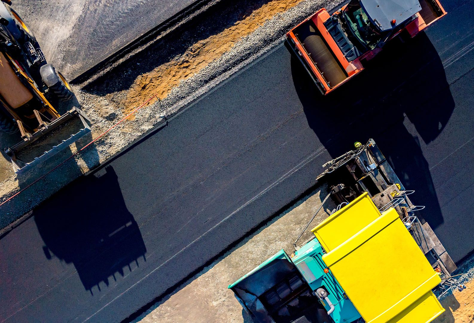 Overhead view of construction vehicles paving a road, including a yellow machine and orange trucks