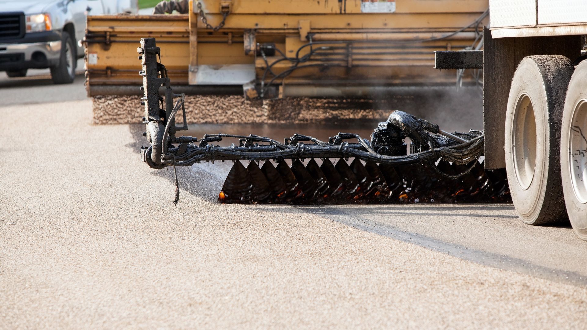 Road grader blade scraping gravel on a construction site, with dust and vehicle wheels visible